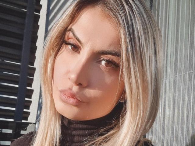 Lellie has blonde hair, brown eyes, and full lips with a slight pout. She's wearing a black turtleneck, and her eyebrows are neatly shaped. Her skin is fair.