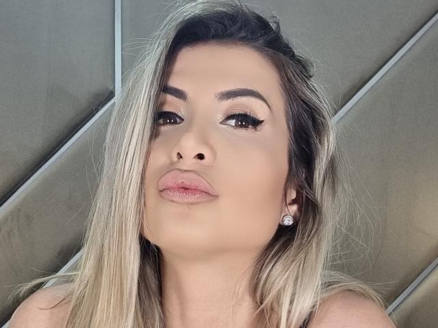 Lellie has blonde hair, thick eyebrows, long eyelashes, pink lipstick, and diamond earrings. Her pouty lips and confident gaze are center. Her face is slightly angled downward.