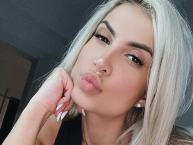 Lellie with pouty lips, blonde hair, and white nails, looking at camera, hand under chin, brown eyes, dark eyebrows, small pearl earrings, soft light on face.