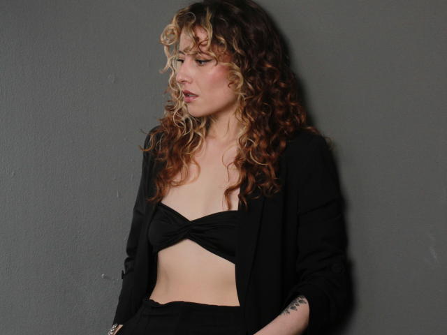VeronicaJoplin with curly blonde and brown hair, wearing a black crop top and jacket, exposing her tits, standing against a gray wall, looking to the side, pouty expression.