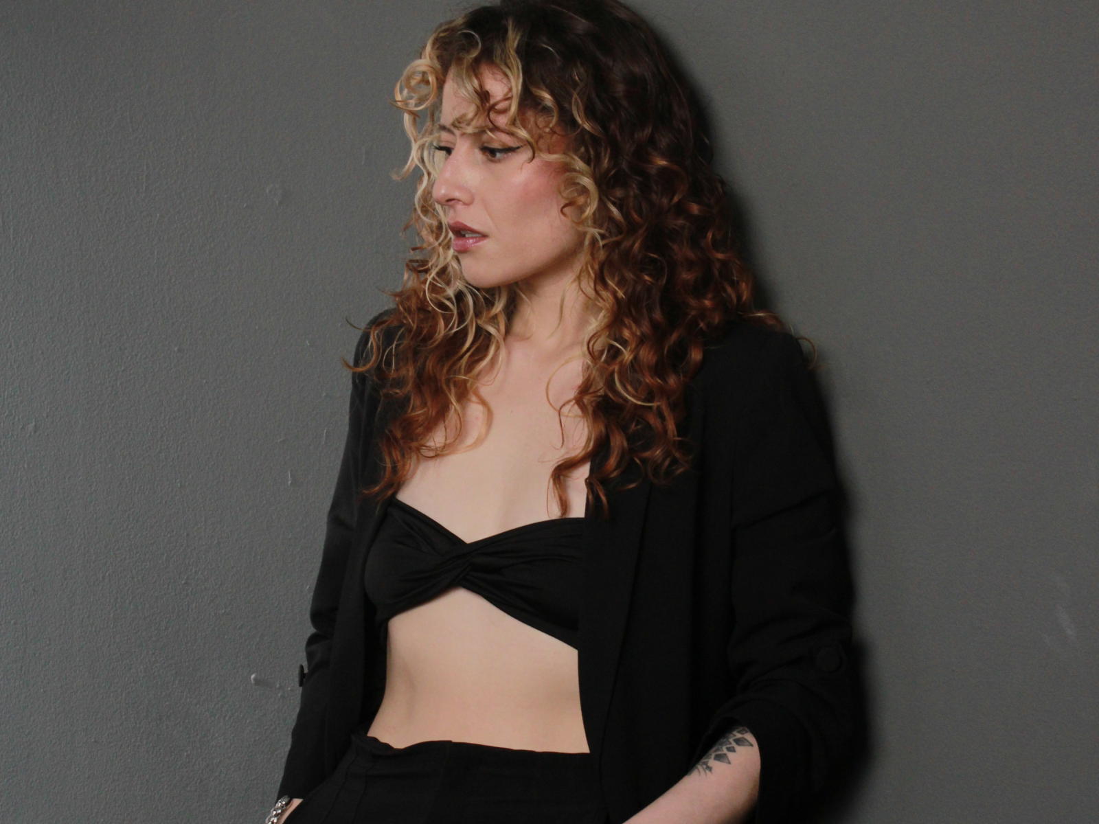 VeronicaJoplin with curly blonde and brown hair, wearing a black crop top and jacket, exposing her tits, standing against a gray wall, looking to the side, pouty expression.