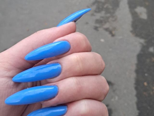 AnnaLuiza's hand with shiny blue, long, pointed acrylic nails, gripping her fingers together. Her nails are glossy and stand out against her pale skin.