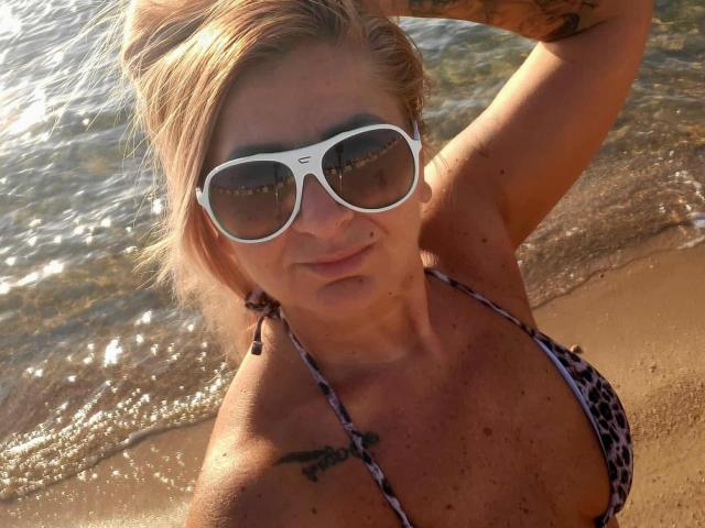 ChaudeEvely with blonde hair, white sunglasses, leopard-print bikini, tattoo on right shoulder, holding hair up, standing at beach with water in background. Boobs are visible, looking at camera.