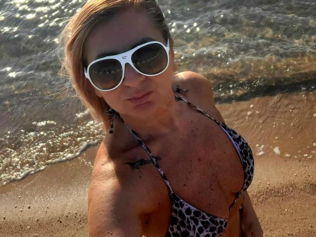 ChaudeEvely with blonde hair, wearing white sunglasses, leopard-print bikini, and small tattoos, standing on a sunlit beach with gentle waves. Boobs visible in the bikini top.
