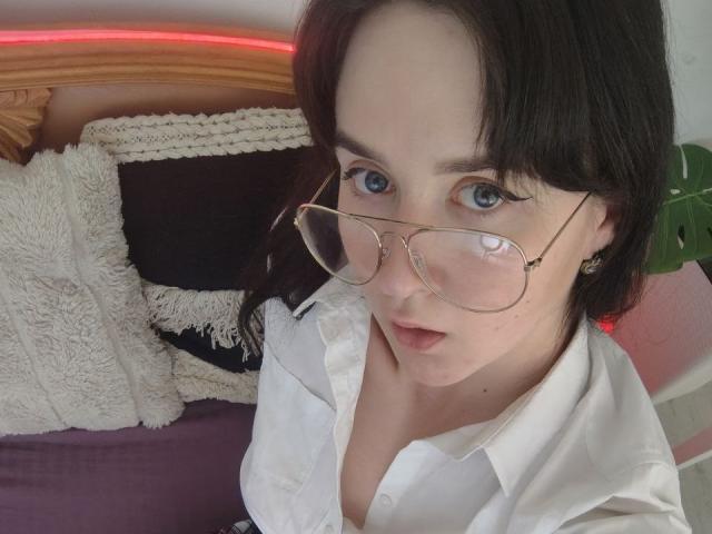 BerryDiva with pale skin, blue eyes, and black hair, wearing glasses and a white shirt, showing her tits slightly, looking up at the camera.
