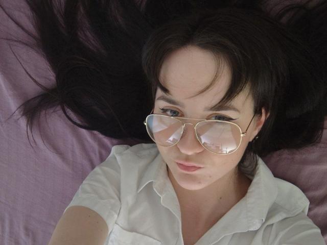 BerryDiva with long black hair, wearing gold aviator sunglasses and a white shirt, lying on a pink bed, showing a serious expression. Her tits are partially visible.