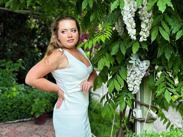 LillSandra with blonde hair in a light blue dress, standing with hands on hips, looking to the side, near a green leafy plant with white flowers.
