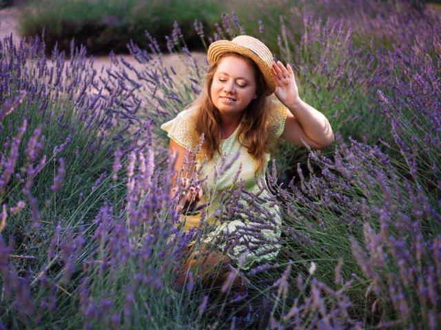 LillSandra with wavy brown hair, wearing a yellow dress and straw hat, sitting in tall purple lavender, eyes closed, hand on hat, in a sunlit field.