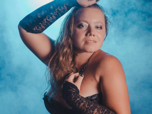 LillSandra with long blonde hair, wearing black lace sleeves and a strapless top, touching her neck, blue background, looking slightly annoyed, showing her tits.