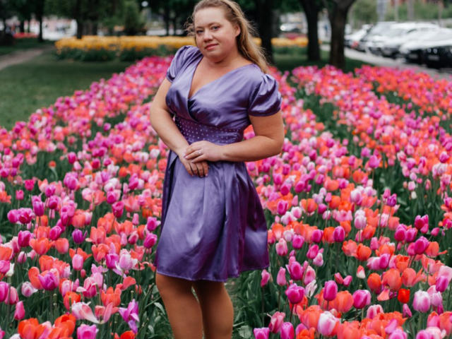 LillSandra stands in a field of pink, red, and purple tulips, wearing a shiny purple dress, hands on her hip, smiling with her blonde hair down.