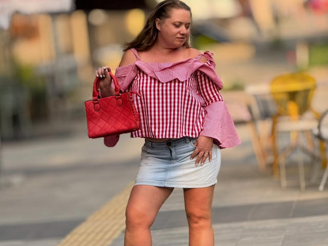 LillSandra with medium tits, wearing a red checkered off-shoulder top and a denim skirt, holds a red quilted handbag, standing outdoors, with blonde hair in a ponytail.