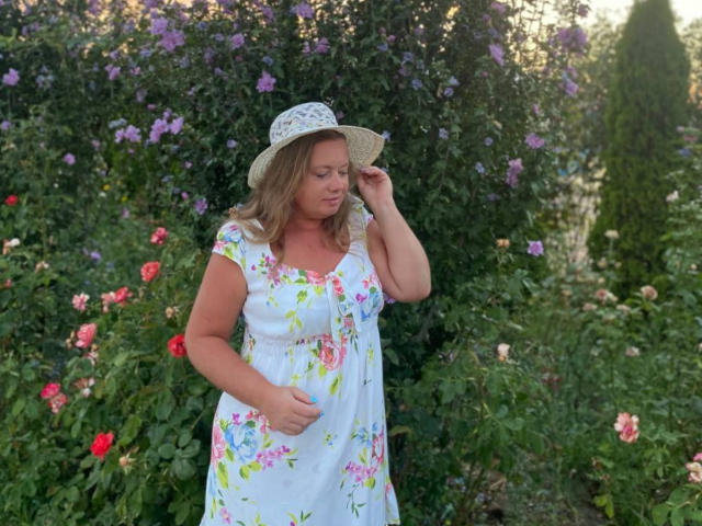 LillSandra, pregnant, wears a floral white dress and straw hat, standing in a garden with red and purple flowers, touching her hat with a gentle smile.