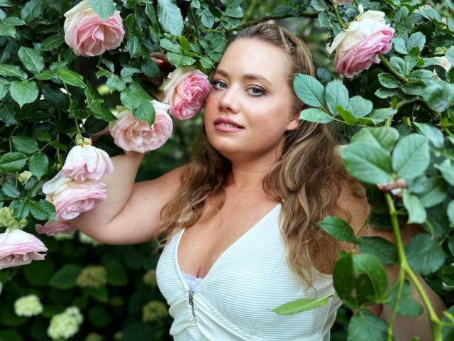 LillSandra with wavy blonde hair, pink roses, green leaves, white dress, and visible boobs, standing amid blooming roses, looking slightly to the side with a soft expression.