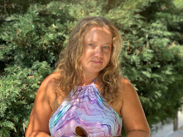 LillSandra with curly blonde hair, wearing a purple and blue wavy-patterned halter top, has a slightly chubby face and is standing in front of green bushes.