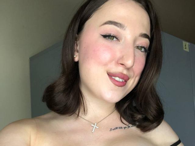 MonAmourR tiene un ligero rubor rosado en las mejillas, delineador de ojos negro y lápiz labial rosa. Está sonriendo, con ojos verdes, cabello castaño de largo medio y un collar con una cruz. Sus pechos son parcialmente visibles..
