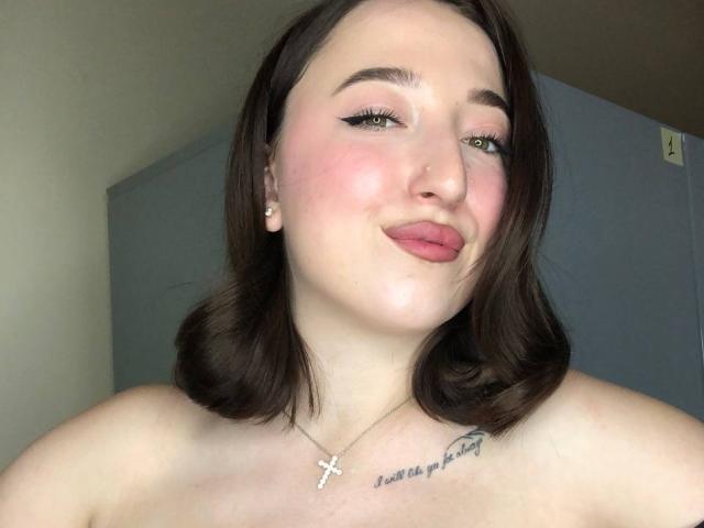 MonAmourR with pink pout, dark brown shoulder-length hair, green eyes, black eyeliner, and a cross necklace, showing a tattoo on the chest, bare shoulders, smiling slightly.
