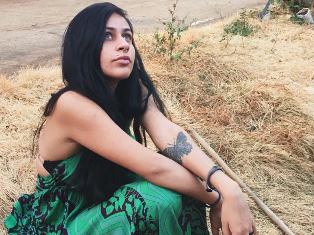 Fendi69 with long black hair, green patterned dress, butterfly tattoo on right arm, sitting on dry hay, holding a wooden stick, looking up with a calm expression.