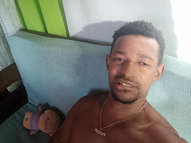 Shirtless Kirilyo with short curly black hair, light brown skin, and a necklace, smiling at the camera. Toy with "MAYBELYN" on it is in the background.