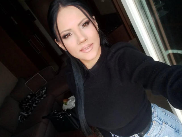 ShadowSirena with long black hair, wearing a black sweater and blue jeans, smiling at the camera, her small tits slightly visible, standing near a glass door.