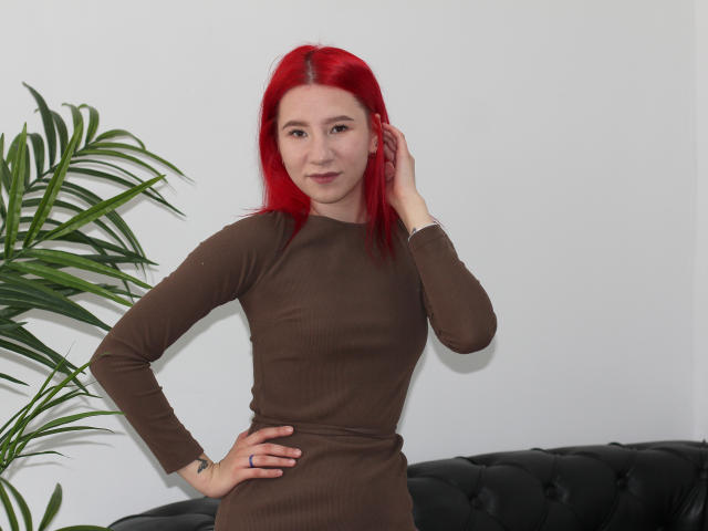 KiraLarkins with red hair, brown long-sleeve dress, hand on hip, touching ear, standing next to green plant, black tufted couch, slight smile, looking at camera.