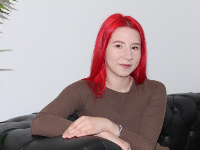 KiraLarkins with red hair, brown shirt, and black leather couch, arms crossed, smiling subtly, white background, wrist bracelet visible.