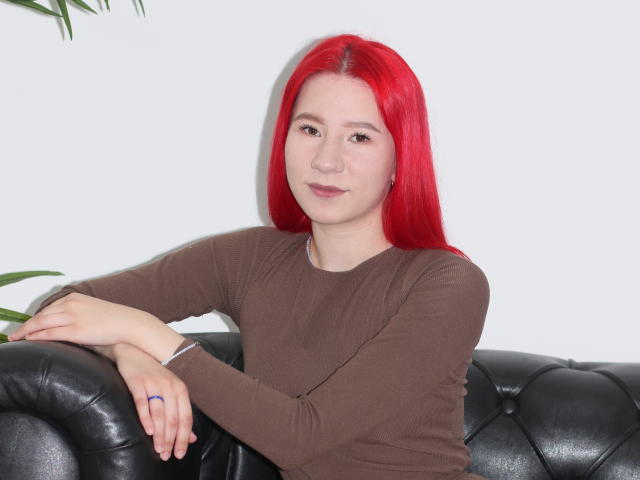 KiraLarkins with bright red hair, brown long-sleeve shirt, black leather couch, and a subtle smile, resting her arms on the couch. She's wearing a blue ring.