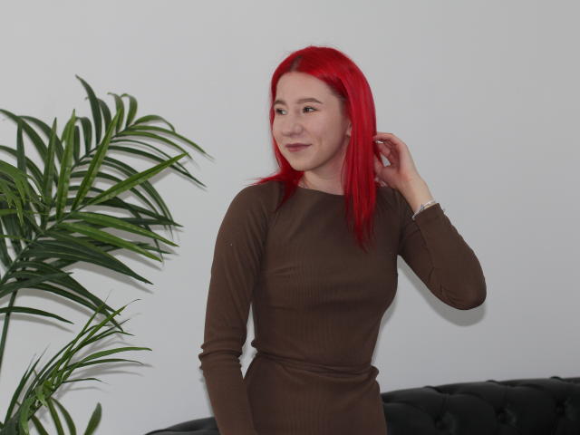 KiraLarkins with bright red hair, wearing a brown long-sleeve dress, smiling, hand in hair, standing next to a green potted plant, black tufted sofa in background.