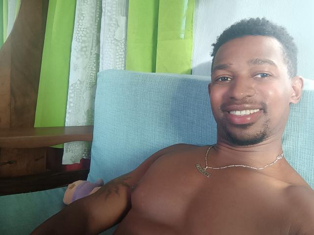 Shirtless Kirilyo with dark skin, short curly hair, and a necklace, smiling, showing a tattoo on his right arm, sitting in a blue chair.