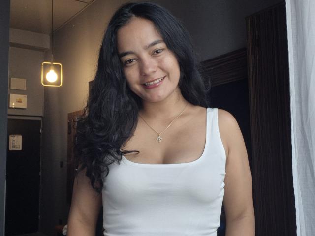LilianaPink with long black wavy hair, smiling, wearing a white tank top that shows her small tits and a gold necklace with a tiny cross. She's standing indoors.