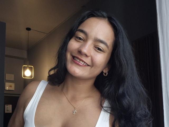 LilianaPink with long black hair, smiling, wearing a white tank top showing her small tits, and a gold necklace with a small pendant. She has a cute face.