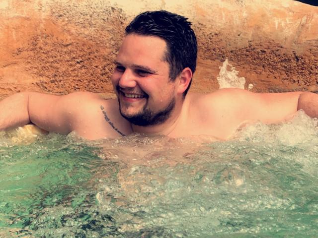 EnergyJumper69, shirtless with short dark hair, beard, and tattoo on right shoulder, smiling in a hot tub with bubbling water. His ass is partially submerged.