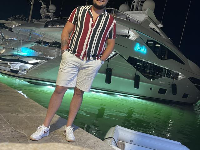 EnergyJumper69 stands in front of a large yacht, wearing a striped shirt, white shorts, and white sneakers, with hands in pockets, green water reflecting under yacht lights.