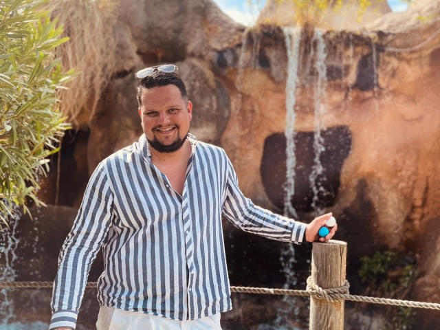 EnergyJumper69, a chubby man with a beard and striped shirt, stands smiling with sunglasses on his head, leaning on a rope, beside a waterfall with his right hand.