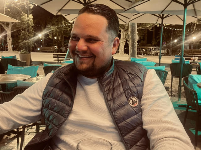 EnergyJumper69, a chubby dude with a dark vest, white shirt, and short beard, smiling at the camera, holding a glass, with a patio and umbrellas in the background.