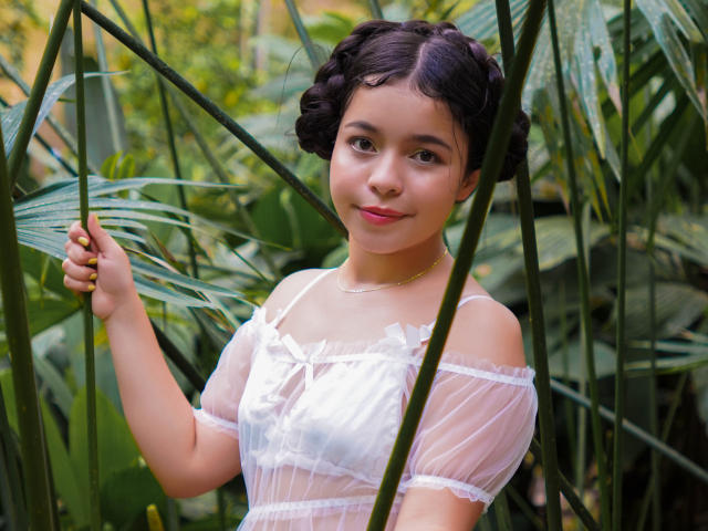 LissyCarter with curly black hair, wearing a sheer white top, standing amidst green leaves, holding a leaf with a gentle smile, pink lips, and a gold necklace.