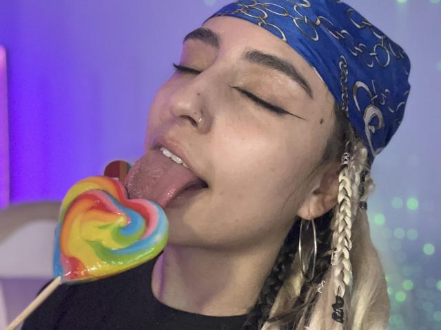 MaveParker with blue bandana, nose ring, and hoop earrings, licking a rainbow heart lollipop, eyes closed, tongue out. Blonde braids, fair skin, and black shirt.