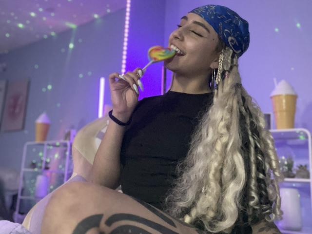 MaveParker with long, curly blonde hair and a blue bandana, sucking a rainbow lollipop, wearing a black top, showing a tattoo on their thigh, in a colorful room.