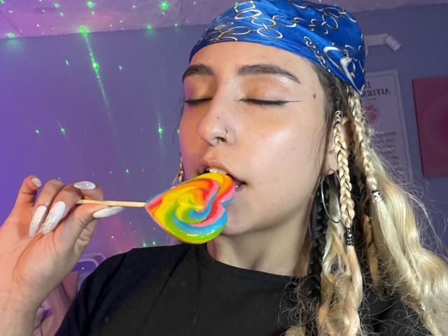 MaveParker with eyes closed, holding a colorful swirl lollipop to her mouth, wearing a blue bandana, black shirt, and braided hair, has white nails and a nose ring.