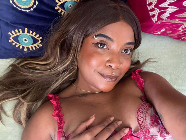 GoddesFox-hot with brown skin, long brown hair, nose piercings, and eye makeup, wearing red lace bra, lying on pillow with blue eyes, smiling, hand on chest.