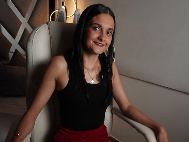 DulceSweety, avec ses cheveux noirs lisses, portant un haut noir sans manches et un pantalon rouge, assise sur une chaise blanche, souriante, avec de petites seins, des hanches de taille moyenne, une peau claire et un collier..