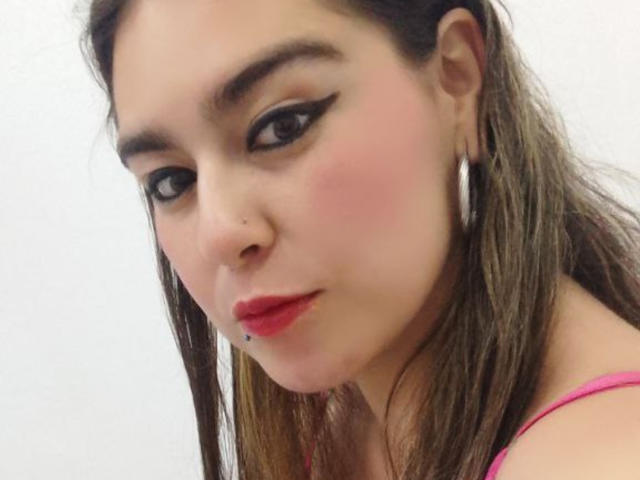 TaylorNova con un leggero rossore sulle guance, eyeliner nero, rossetto rosso, capelli castani, orecchini a cerchio argentati e un piercing sul labbro inferiore, che guarda in basso con un leggero sorriso..