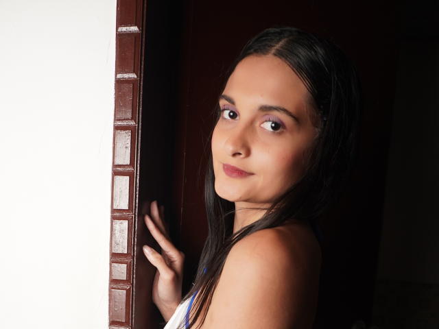 DulceSweety, avec ses longs cheveux noirs, sa peau brun clair et ses petites seins, regardant par-dessus son épaule, debout près d'une porte avec un cadre marron, et un léger sourire sur son visage..