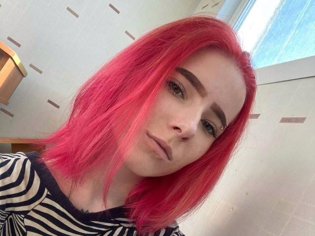 RosyLight ha i capelli rosa acceso, un piercing al naso e indossa una camicia a righe, guardando la fotocamera con un'espressione leggermente corrucciata. Le sue sopracciglia sono folte e scure..