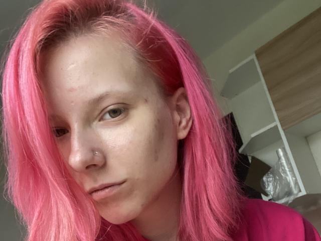 RosyLight tiene el pelo rosa, un piercing en la nariz, ojos verdes y un pequeño grano en la mejilla, lleva una camisa rosa y mira a la cámara con una expresión neutra..