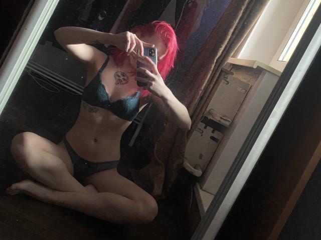 RosyLight, con i capelli rossi e lucidi, indossa un reggiseno e una biancheria intima in pizzo verde scuro, è seduta a gambe incrociate e sta scattando un selfie allo specchio, mostrando i suoi seni e i suoi genitali, in una stanza poco illuminata..