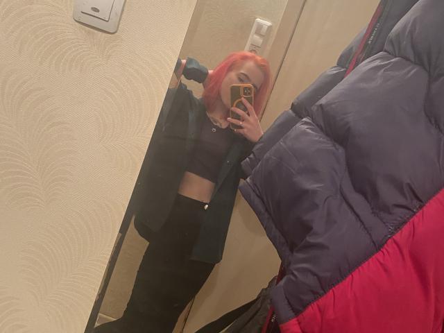 RosyLight, con el cabello rojo brillante, vestida con un top corto negro y pantalones negros, se toma un selfie frente al espejo, mostrando una chaqueta azul y roja hinchada en primer plano..