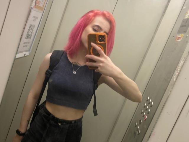 RosyLight con i capelli rosa, top corto scuro senza maniche, pantaloni neri a vita alta, che tiene un telefono arancione, selfie allo specchio, seno piccolo, magra, collana d'argento, zaino nero, ascensore, selfie..