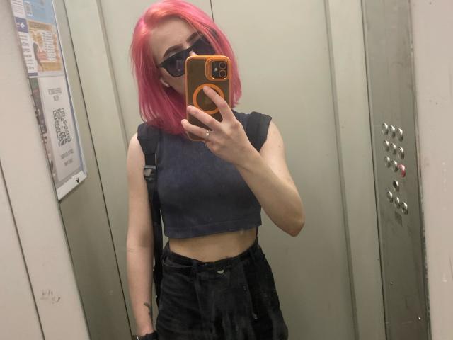 RosyLight mit rosa Haaren, einem schwarzen Crop-Top, schwarzen, hochgeschnittenen Hosen, einer schwarzen Maske und Sonnenbrille, die ein Selfie vor einem Spiegel in einem Aufzug macht und ein orangefarbenes Handy hält..