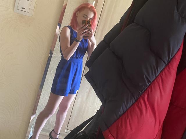 RosyLight, avec ses cheveux roses, portant une robe bleue, prenant un selfie devant un miroir, montrant ses fesses et ses jambes, tenant un téléphone, avec une veste matelassée noire et rouge au premier plan..