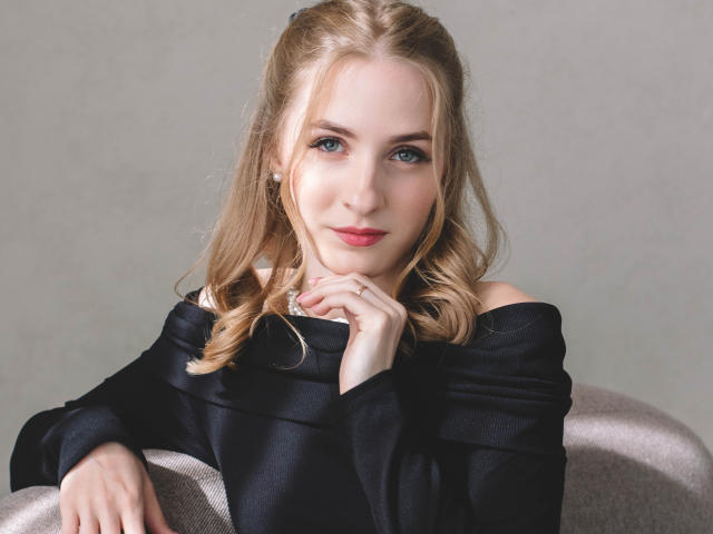 EvaDaly with blonde hair, blue eyes, and pink lips, wearing a black off-shoulder top, sitting on a gray chair, hands near her face, looking directly at the camera.